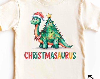 May include: A white vintage t-shirt with a green dinosaur wearing a Santa hat and holding a Christmas tree with lights. The text "CHRISTMASAURUS" is printed in red, yellow, and green letters on the shirt.