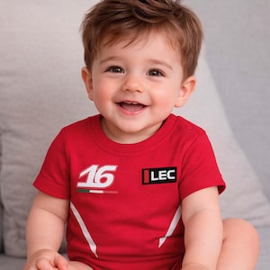 Könnte beinhalten: Ein lächelndes Baby trägt einen roten Body mit der Nummer "16" und den Buchstaben "LEC" auf der Vorderseite. Der Body hat weiße Akzente und ein kleines italienisches Flaggen-Design. Das Baby sitzt auf einem grauen Sofa.
