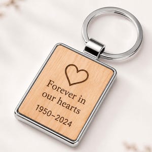 May include: A rectangular keyring featuring a light wood inlay set within a polished silver-tone metal frame. The wood surface is engraved with a heart symbol above the text "Forever in our hearts" and the years "1950-2024" in a dark font.