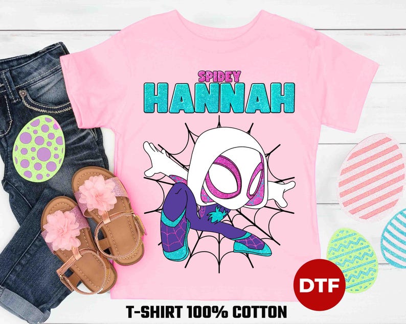 May include: Pink t-shirt with a cartoon image of Spider-Gwen, a female superhero with white and black costume, and the text "SPIDEY HANNAH" in blue and green glitter letters. The shirt is made of 100% cotton and has a DTF print.