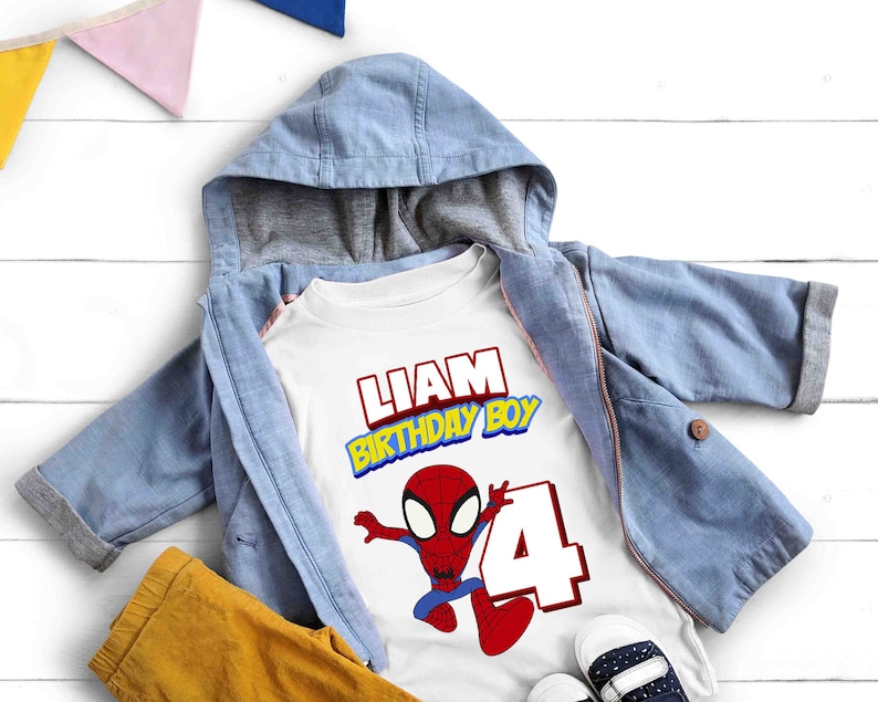 May include: White t-shirt with a red and blue Spiderman graphic, the text "Liam Birthday Boy" and the number "4". The shirt is on top of a blue denim jacket and yellow pants.