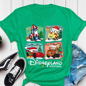 May include: Green t-shirt with a Christmas themed design featuring characters from the Disney Pixar movie Cars. The design includes Lightning McQueen, Mater, and other characters wearing Santa hats and driving decorated cars. The text "Disneyland Christmas" is printed below the design.