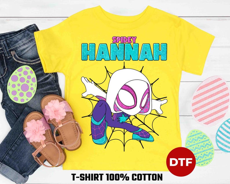 May include: Yellow t-shirt with a cartoon image of Spider-Gwen, a female superhero with white skin, a black and purple suit, and a spider web design. The text "SPIDEY HANNAH" is printed on the shirt.