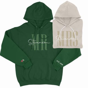 Puede incluir: Un conjunto a juego de sudaderas con capucha verde y beige con las palabras "MR Stevenson" y "MRS Stevenson" impresas en ellas. La sudadera con capucha verde tiene un corazón rojo y el texto "EST. 2020" en las mangas.