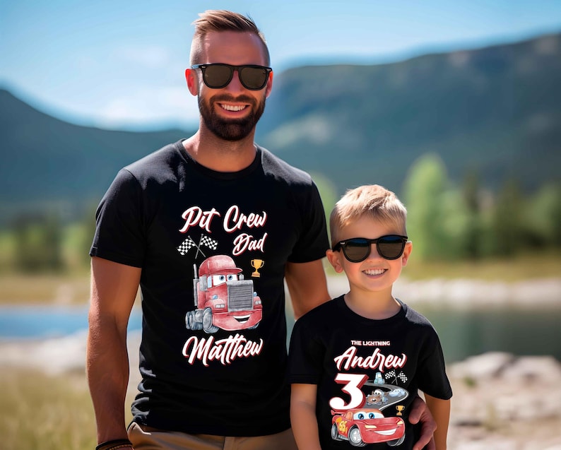 May include: Two black t-shirts with a Cars theme. The adult shirt says "Pit Crew Dad Matthew" with a red race car truck. The child's shirt says "The Lightning Andrew 3" with a red race car.