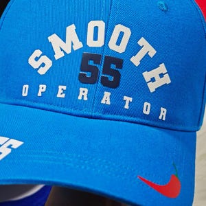 May include: A blue baseball cap with white text that reads "SMOOTH 55 OPERATOR" and a red chili pepper graphic. The number 55 is in navy blue.