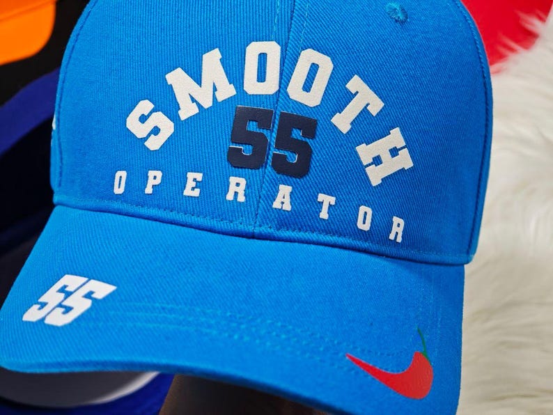 May include: A blue baseball cap with white text that reads "SMOOTH 55 OPERATOR" and a red chili pepper graphic. The number 55 is in navy blue.