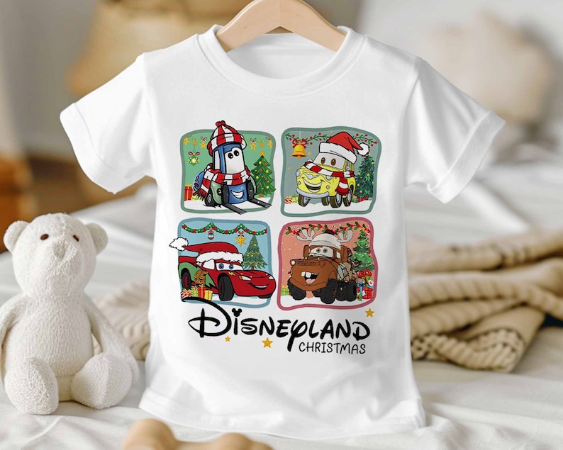 May include: White t-shirt with a Christmas themed design featuring four characters from the Disney Pixar movie Cars. The characters are Mater, Lightning McQueen, Tow Mater, and a blue car wearing a Santa hat. The text "Disneyland Christmas" is printed below the characters.