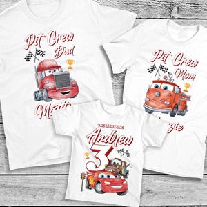 May include: Three white t-shirts with a Cars theme. The shirts feature a red race car, a red fire truck, and a red semi-truck. The text on the shirts reads "Pit Crew Dad", "Pit Crew Mom", and "The Lightning Andrew 3".