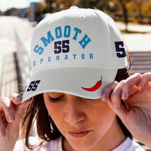 May include: White baseball cap with blue and black text that reads "SMOOTH 55 OPERATOR 5" and a red chili pepper graphic. The number 55 is also on the side of the cap.