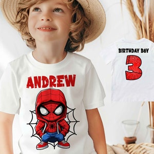 May include: White t-shirt with a red and blue cartoon Spiderman design and the name "ANDREW" printed in red. The shirt is being worn by a child wearing a straw hat.