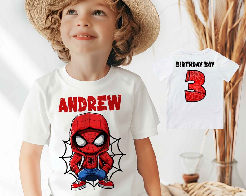 May include: White t-shirt with a red and blue cartoon Spiderman design and the name "ANDREW" printed in red. The shirt is being worn by a child wearing a straw hat.