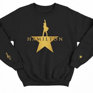 Peut inclure: Sweat-shirt noir avec une étoile dorée et le mot "HAMILTON" imprimé sur le devant. L'étoile a une silhouette d'une personne à l'intérieur.