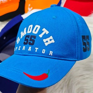 May include: A blue baseball cap with a white logo that says "SMOOTH 55 OPERATOR" and a red chili pepper design on the front. The number 55 is also printed on the side of the cap.