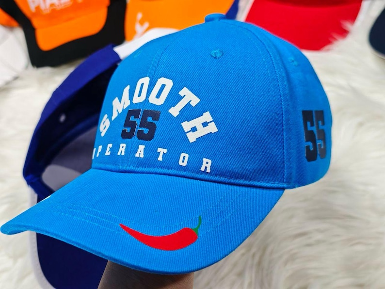 May include: A blue baseball cap with a white logo that says "SMOOTH 55 OPERATOR" and a red chili pepper design on the front. The number 55 is also printed on the side of the cap.
