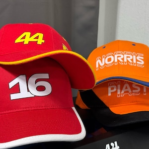 Op de afbeelding: Twee baseballpetten, een rode met witte nummers "16" en "44" en een gele klep, de andere oranje met witte tekst "NORRIS" en "PIASTRI" en een blauwe klep.