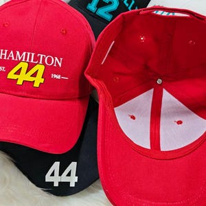 May include: Red baseball cap with a black brim and a white logo that says "HAMILTON -EST. 44 1968- ". The number 44 is in yellow. The inside of the cap is white.
