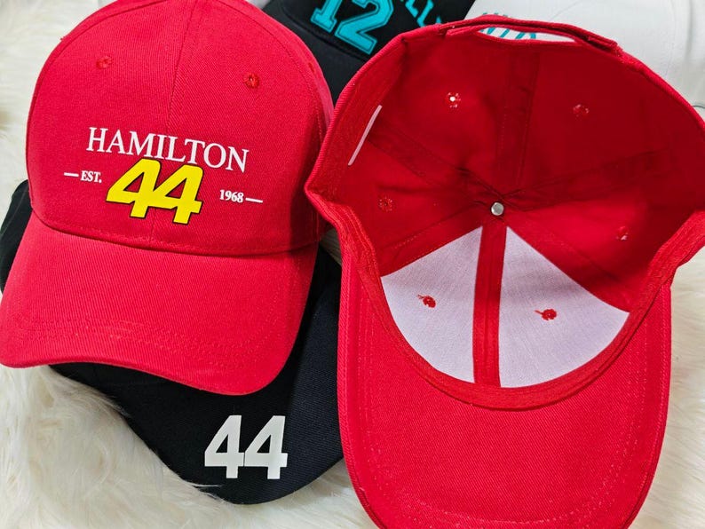 May include: Red baseball cap with a black brim and a white logo that says "HAMILTON -EST. 44 1968- ". The number 44 is in yellow. The inside of the cap is white.