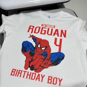 Op de afbeelding: Wit T-shirt met een rode en blauwe Spiderman-afbeelding, de tekst "The Amazing Roguan 4 Birthday Boy" in rood.