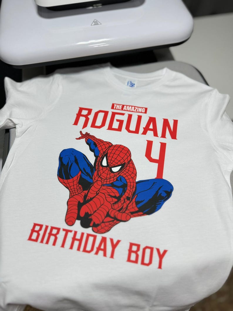 Op de afbeelding: Wit T-shirt met een rode en blauwe Spiderman-afbeelding, de tekst "The Amazing Roguan 4 Birthday Boy" in rood.
