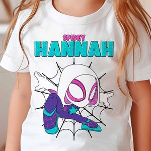 May include: White t-shirt with a graphic of Spider-Gwen, a female superhero, in a white and purple suit. The text "SPIDEY HANNAH" is printed on the shirt in turquoise glitter.