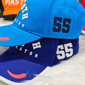 May include: Two baseball caps, one blue and one dark blue, with white stitching and a red chili pepper design. The blue cap has the text "OTH RATOR" and the number "55" in navy blue. The dark blue cap has the text "RATOR" and the number "55" in white.