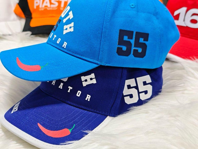 May include: Two baseball caps, one blue and one dark blue, with white stitching and a red chili pepper design. The blue cap has the text "OTH RATOR" and the number "55" in navy blue. The dark blue cap has the text "RATOR" and the number "55" in white.