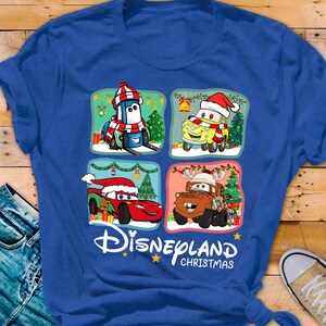 May include: Blue t-shirt with a Christmas themed design featuring characters from the Disney Pixar movie Cars. The design includes Lightning McQueen, Mater, and Tow Mater wearing Santa hats and surrounded by Christmas decorations. The text "Disneyland Christmas" is printed below the design.