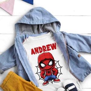 May include: White t-shirt with a cartoon image of Spiderman wearing a red and blue suit and a red hooded sweatshirt. The text "ANDREW" is printed in red above the image.