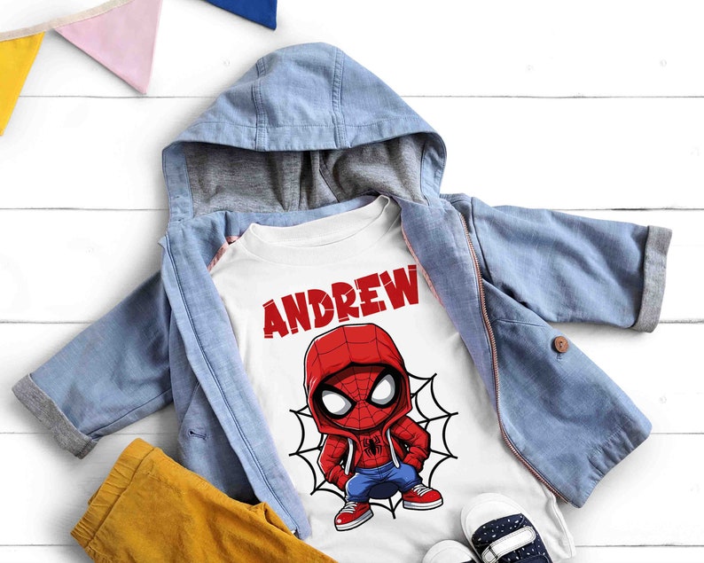 May include: White t-shirt with a cartoon image of Spiderman wearing a red and blue suit and a red hooded sweatshirt. The text "ANDREW" is printed in red above the image.