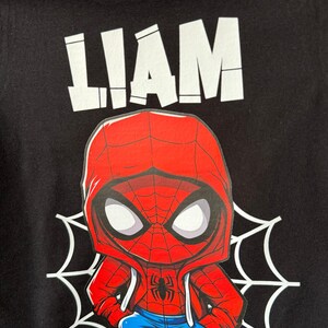 May include: Black t-shirt with a white graphic of the name "LIAM" and a cartoon image of Spiderman wearing a red and blue hoodie, blue jeans, and red sneakers. The Spiderman graphic is on a white spiderweb.