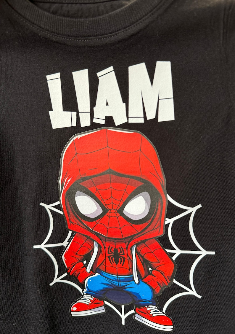 May include: Black t-shirt with a white graphic of the name "LIAM" and a cartoon image of Spiderman wearing a red and blue hoodie, blue jeans, and red sneakers. The Spiderman graphic is on a white spiderweb.