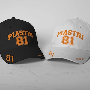 Puede incluir: Dos gorras de béisbol, una negra y una blanca, con el nombre "PIASTRI" y el número "81" en naranja en la parte delantera. La gorra negra tiene una pequeña bandera australiana en el lateral.