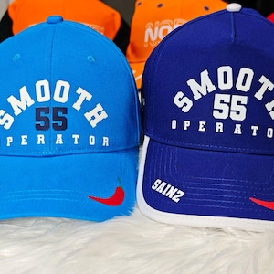 Może przedstawiać: Dwie czapki bejsbolowe, jedna niebieska, a druga jasnoniebieska, z napisem "SMOOTH 55 OPERATOR" z przodu. Obie czapki mają po bokach biały wzór papryczki chili.