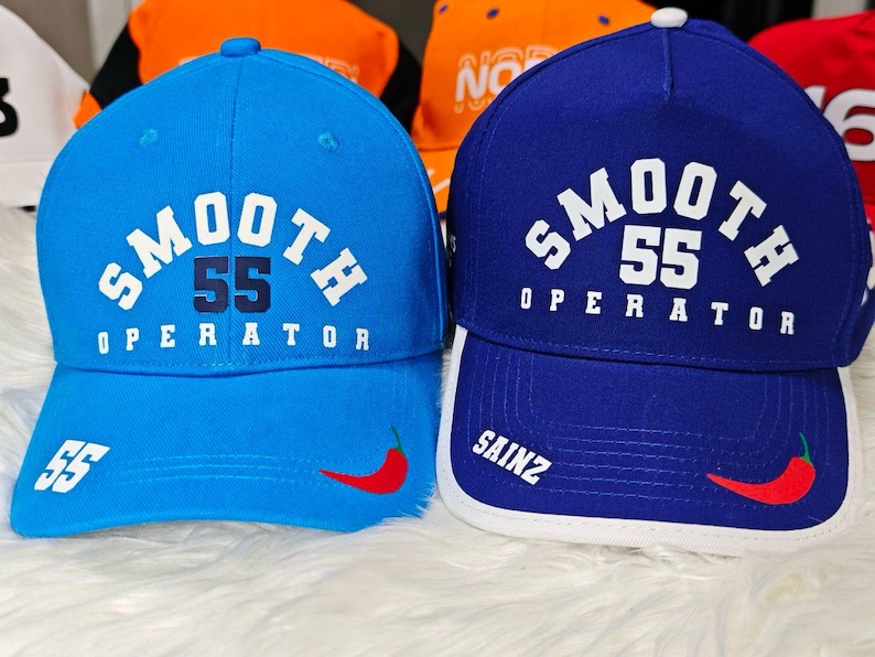 May include: Two baseball caps, one blue and one light blue, with the text "SMOOTH 55 OPERATOR" on the front. Both caps have a white chili pepper design on the side.