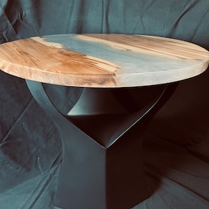 Puede incluir: Mesa auxiliar redonda con tapa de madera y base de metal negro. La tapa presenta un patrón de vetas de madera natural con un río de resina epoxi transparente. La base tiene un diseño geométrico.