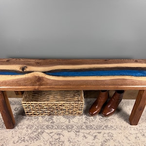 May include: A wooden bench with a blue and black epoxy resin inlay. The bench has two legs and is sitting on a rug. There is a wicker basket and a pair of brown boots under the bench.