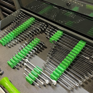 May include: A collection of shiny, silver wrenches neatly organized in a gray toolbox drawer. The wrenches are held in place by bright green holders. A green handled tool is in the foreground.