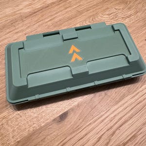 May include: A rectangular, olive-green storage container with a hinged lid. The lid features an orange arrow design. The container is made of a durable material and is designed for secure storage. The container is sitting on a wooden surface.