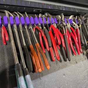 May include: A collection of hand tools, including pliers and other metal tools, neatly organized in a drawer. The tools have red, black, and orange handles, and are held in place by purple tool holders. The drawer is black.
