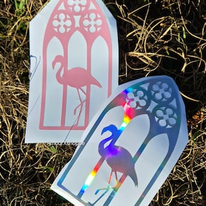 May include: Two flamingo-themed stickers. One is pink, the other is holographic with rainbow hues. Both stickers feature a flamingo silhouette within a gothic arch design. The stickers are on a white background.