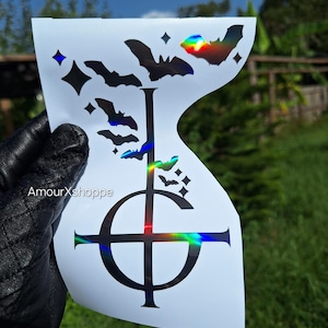 May include: White sticker featuring a black inverted cross and bat design. The sticker has a holographic effect, with bats and diamond shapes. The text "AmourXshoppe" is visible on the sticker.