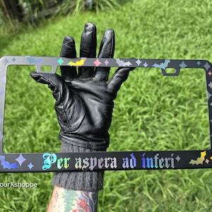 May include: A black license plate frame featuring a holographic design of small bats and four-pointed stars. The bottom edge displays the text "Per aspera ad inferi" in a gothic font, also rendered in a holographic finish that reflects light.