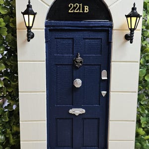 Puede incluir: Una r&eacute;plica en miniatura de la ic&oacute;nica puerta del 221B Baker Street, con una puerta azul, un marco de puerta negro y letras doradas. La puerta tiene un llamador negro y una manija de puerta plateada. La puerta est&aacute; colocada contra una pared blanca con farolas negras a cada lado.