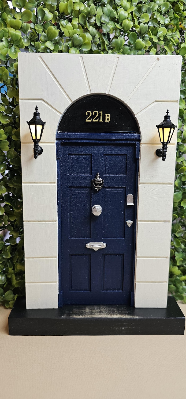 Puede incluir: Una r&eacute;plica en miniatura de la ic&oacute;nica puerta del 221B Baker Street, con una puerta azul, un marco de puerta negro y letras doradas. La puerta tiene un llamador negro y una manija de puerta plateada. La puerta est&aacute; colocada contra una pared blanca con farolas negras a cada lado.