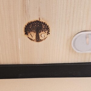 Puede incluir: Una caja de madera con una base negra. La caja tiene un dise&ntilde;o de &aacute;rbol quemado en madera con las letras "VG" dentro del &aacute;rbol. La caja tiene un cierre de pl&aacute;stico transparente en el lateral.