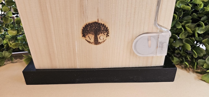 Puede incluir: Una caja de madera con una base negra. La caja tiene un dise&ntilde;o de &aacute;rbol quemado en madera con las letras "VG" dentro del &aacute;rbol. La caja tiene un cierre de pl&aacute;stico transparente en el lateral.
