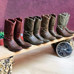 Può includere: Una collezione di stivali da cowboy in miniatura in varie tonalità di marrone e verde, disposti su una tavola di legno. Gli stivali sono esposti su uno sfondo viola, con una moneta visibile in primo piano. Gli stivali sono una deliziosa rappresentazione dell'arredamento a tema western.
