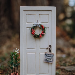 May include: A miniature white door with a green wreath decorated with red berries. A sign hangs on the door that reads "J'habite chez mon CHAT".