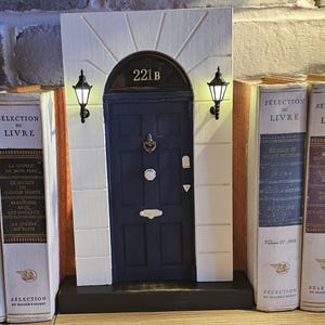 Puede incluir: Una r&eacute;plica en miniatura de una casa adosada de Londres con una puerta azul y el n&uacute;mero 221B sobre la puerta. La casa adosada est&aacute; flanqueada por dos sujetalibros con el texto "Selection du Livre" en los lomos.